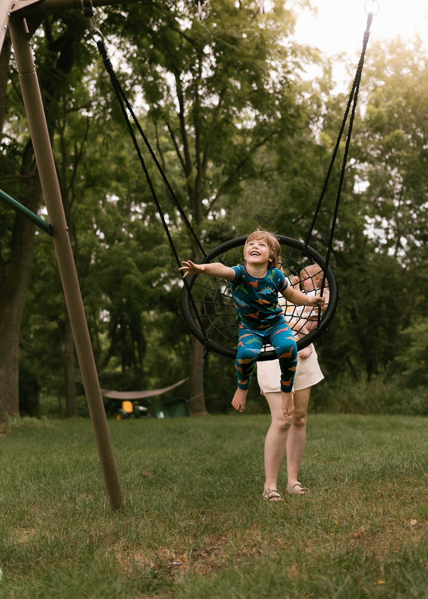 A child enjoys swinging on a playground swing set in a wooded area during golden hour lighting.