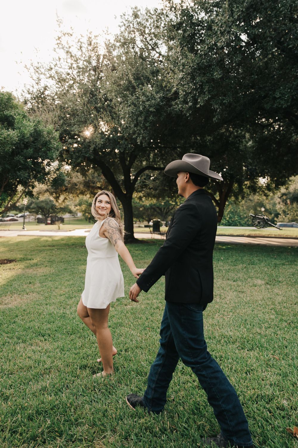 A couple holds hands and playfully spins in a grassy park during a sunny afternoon photo session.