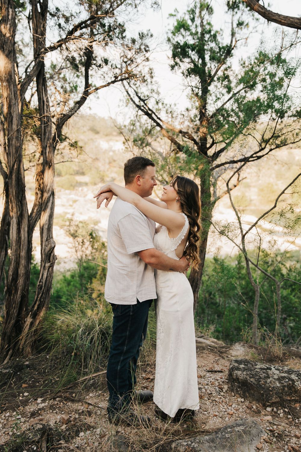 A romantic couple embraces on a nature trail surrounded by trees and golden afternoon light.