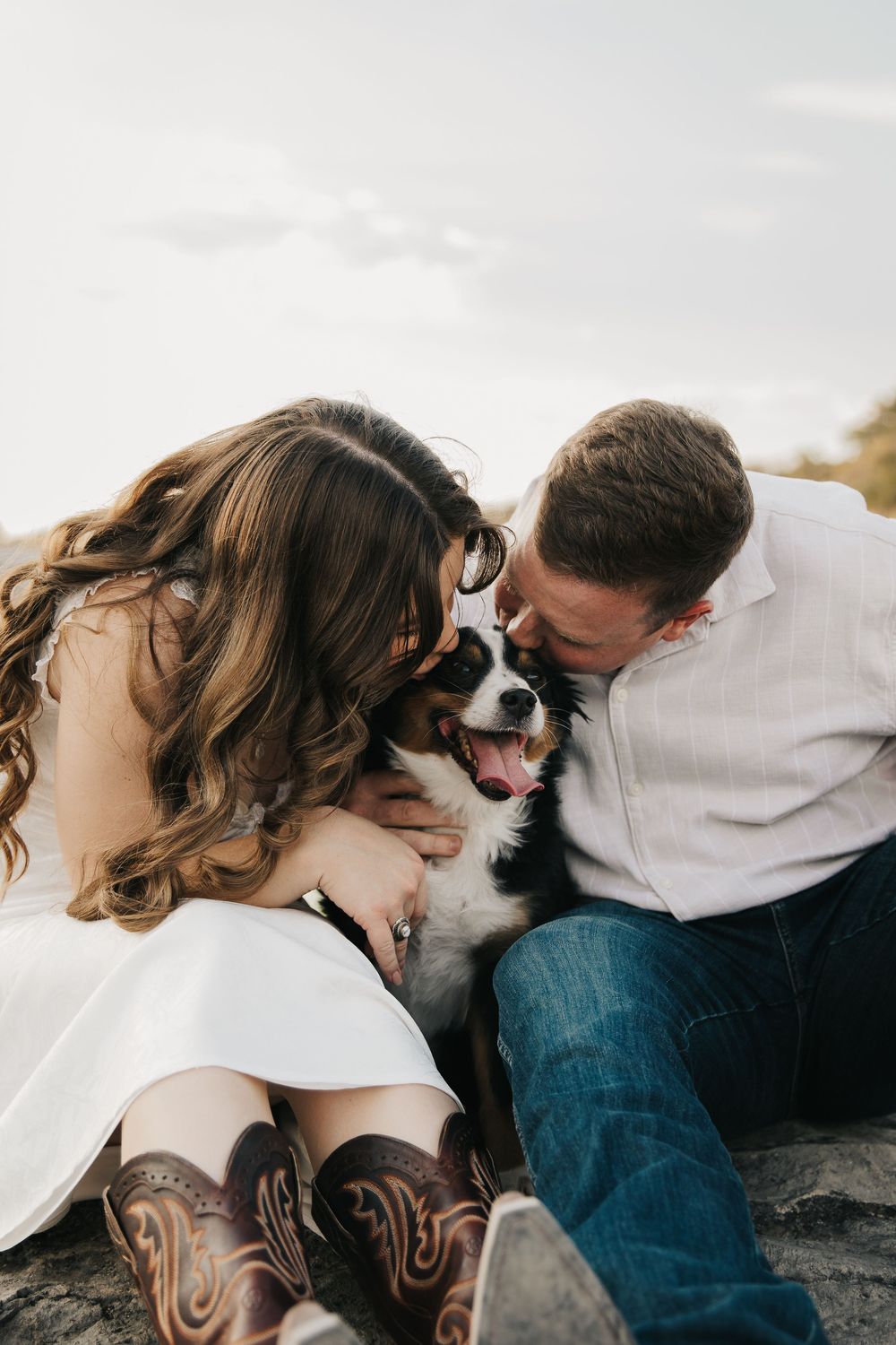 A couple in casual attire shares tender moments with their dog while sitting outdoors in a natural setting.