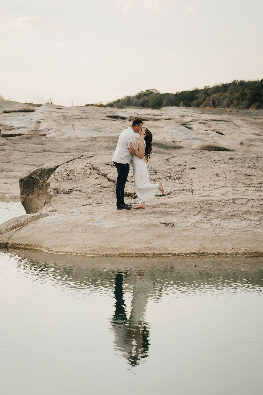A romantic couple embraces on a rocky coastal cliff with their reflection visible in the water below during sunset.