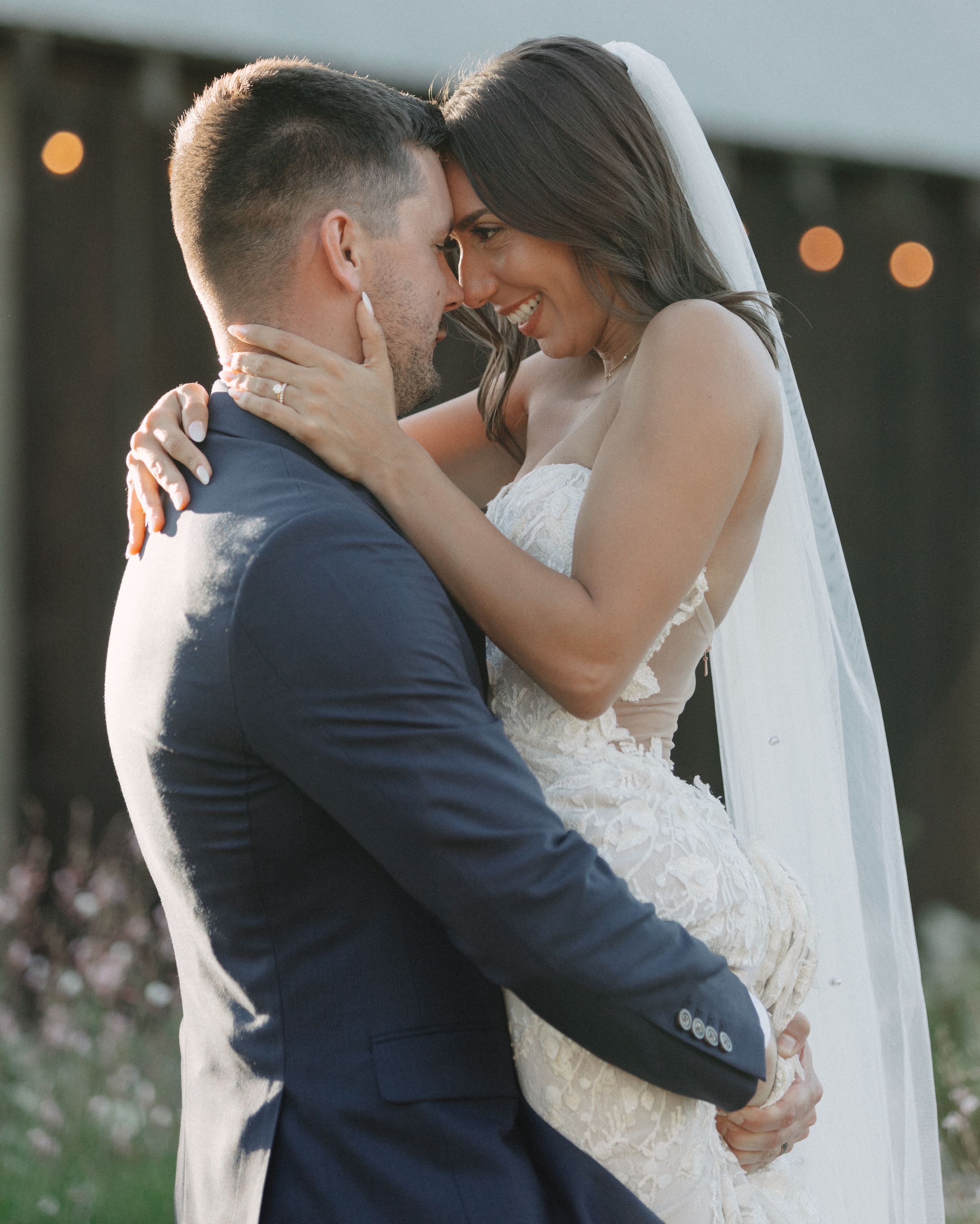 A romantic moment captured between newlyweds embracing outdoors with soft bokeh lights in the background.
