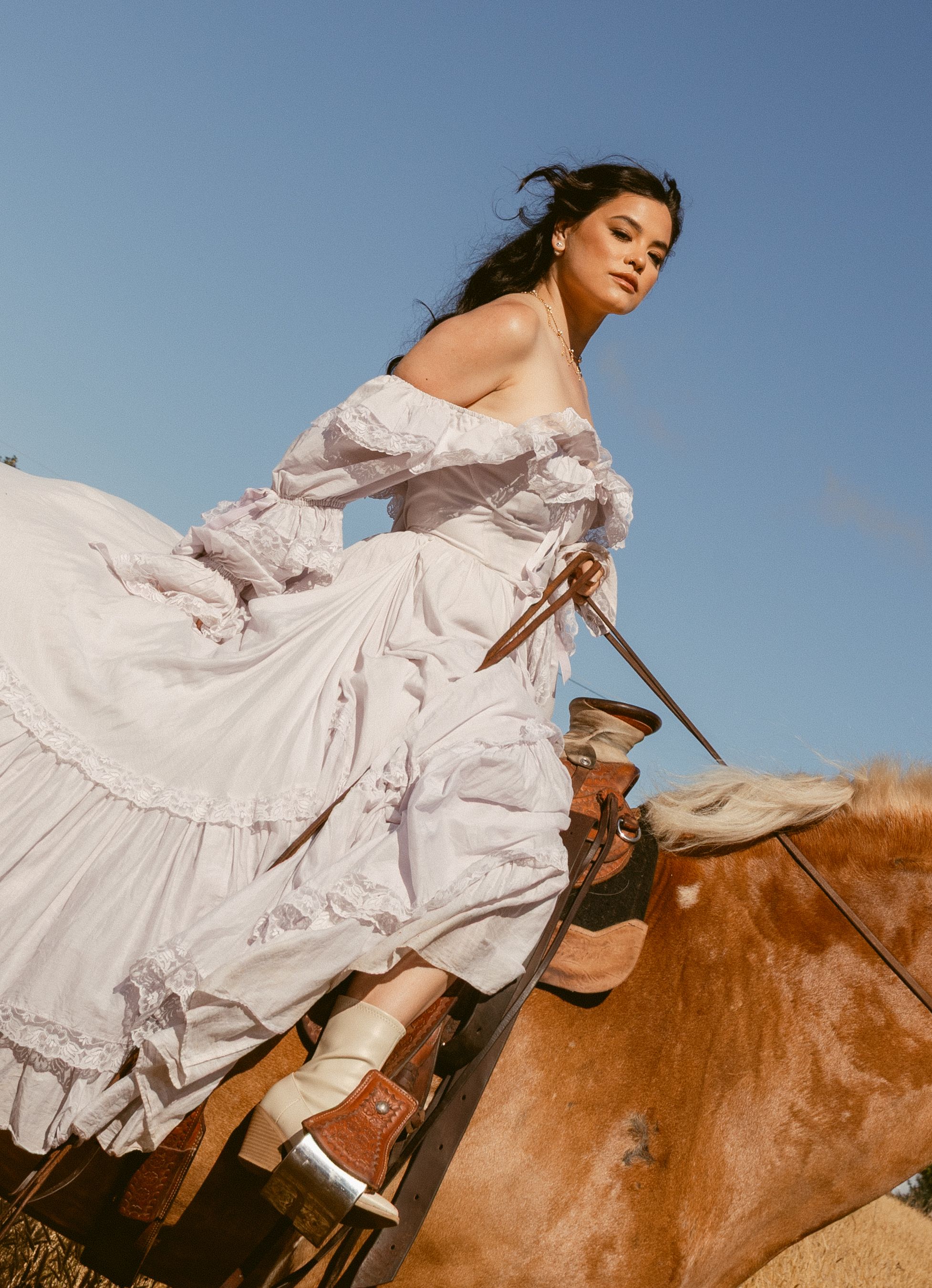 A figure in a flowing white dress rides a brown horse against a blue sky backdrop in a dreamy outdoor photoshoot.