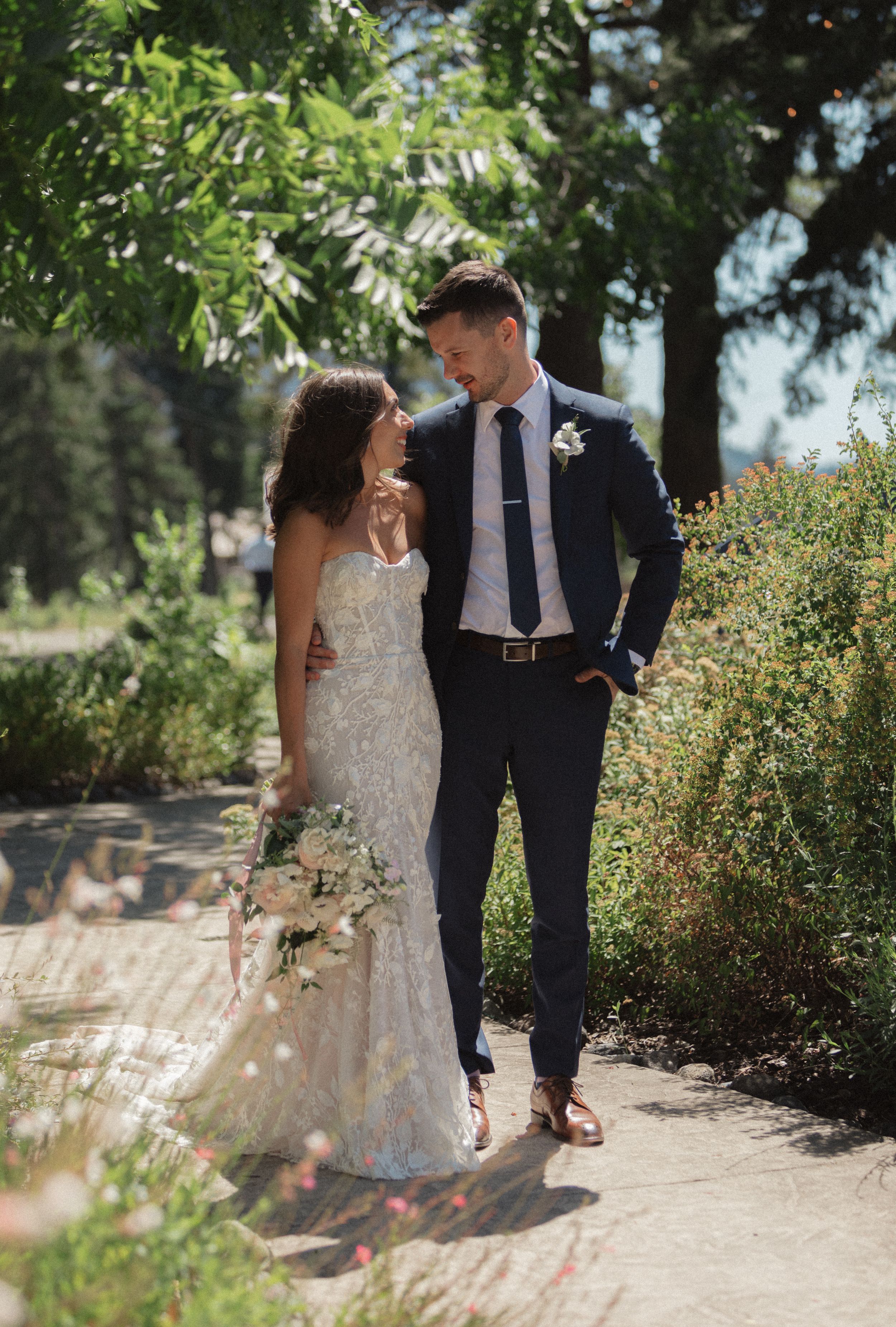 A couple in wedding attire walks together on a garden path surrounded by lush greenery on a sunny day.