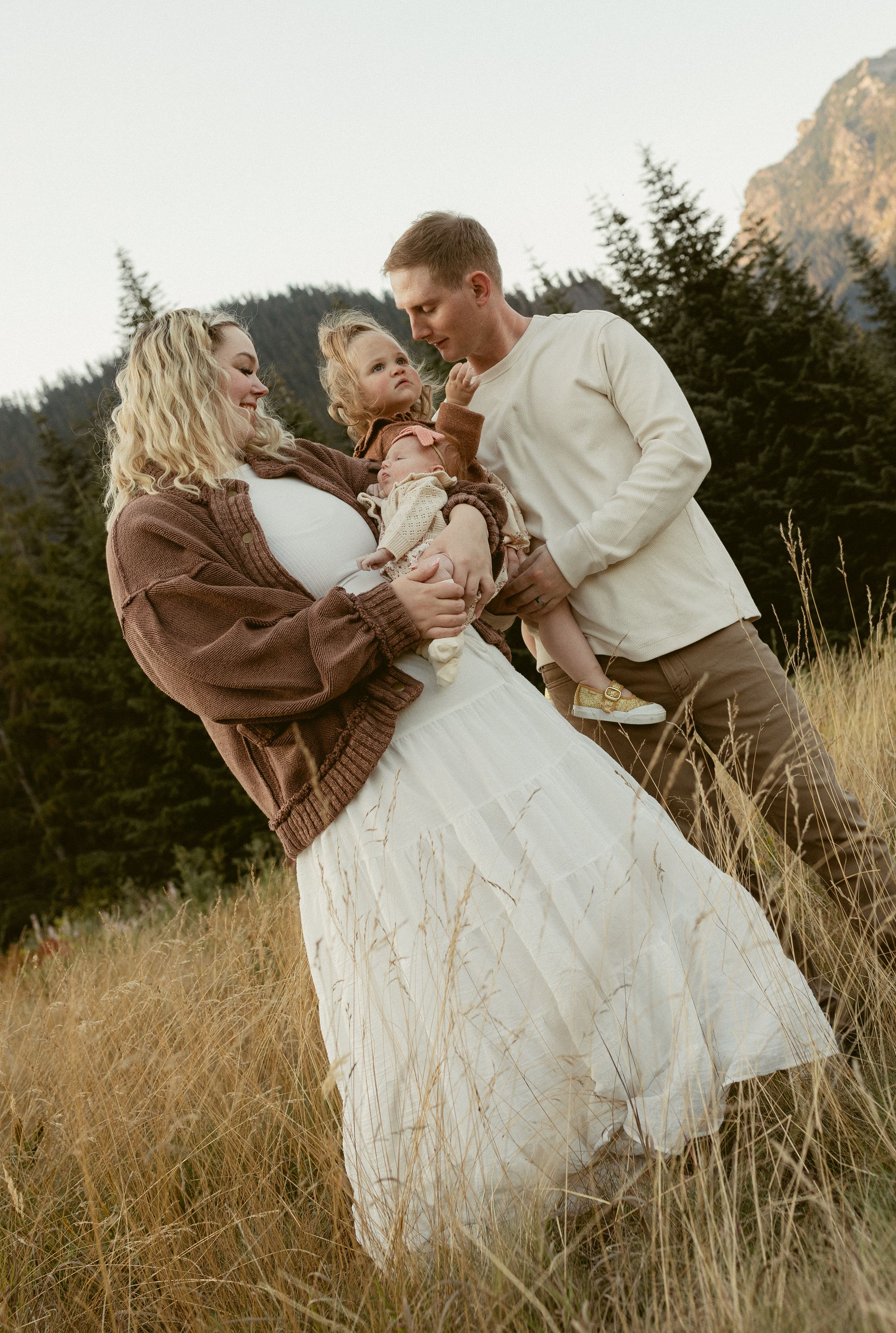 Romantic couple embraces in golden field with mountain backdrop during sunset photoshoot.