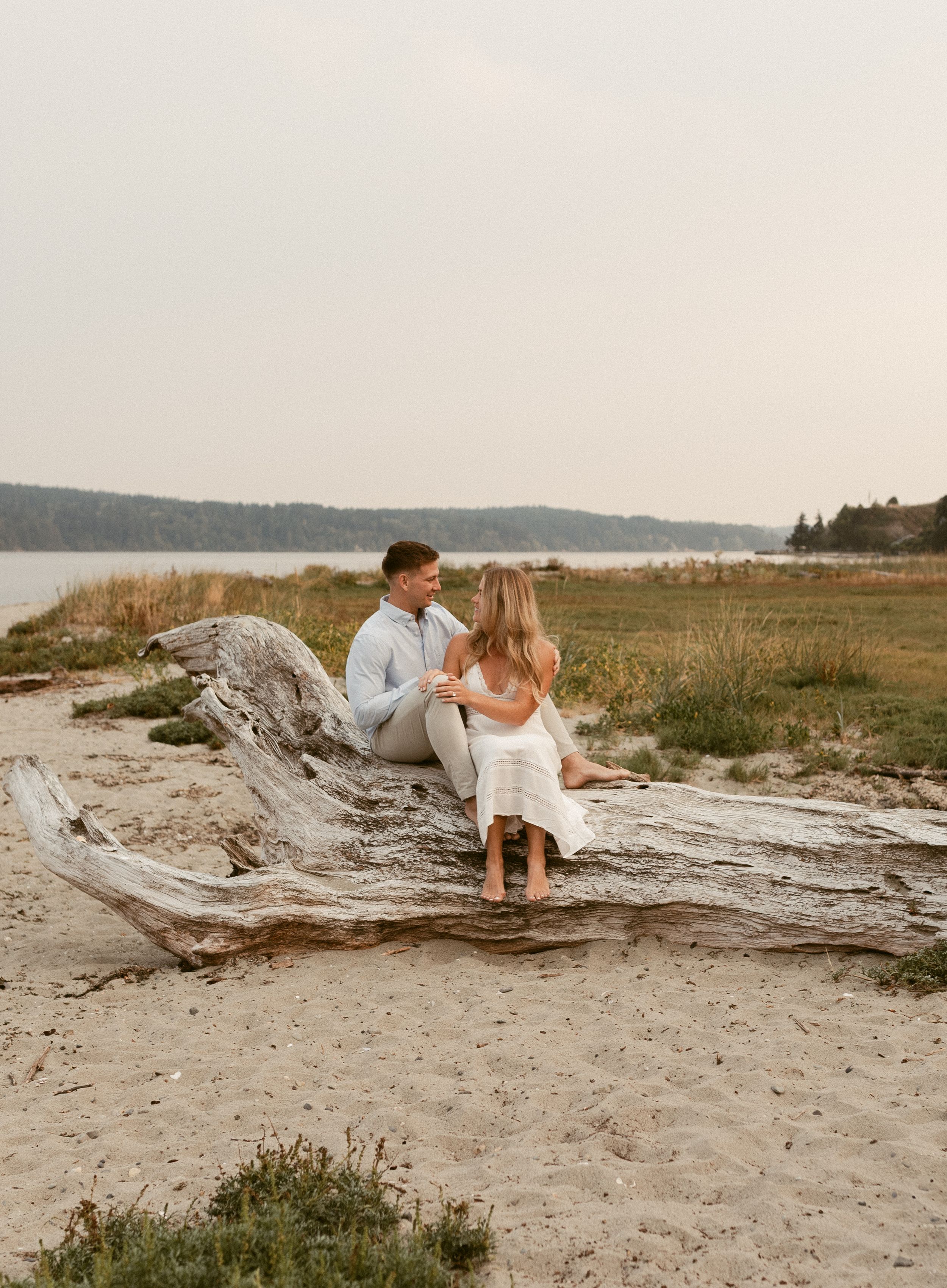 A couple shares an intimate moment on driftwood along a peaceful beach at sunset.