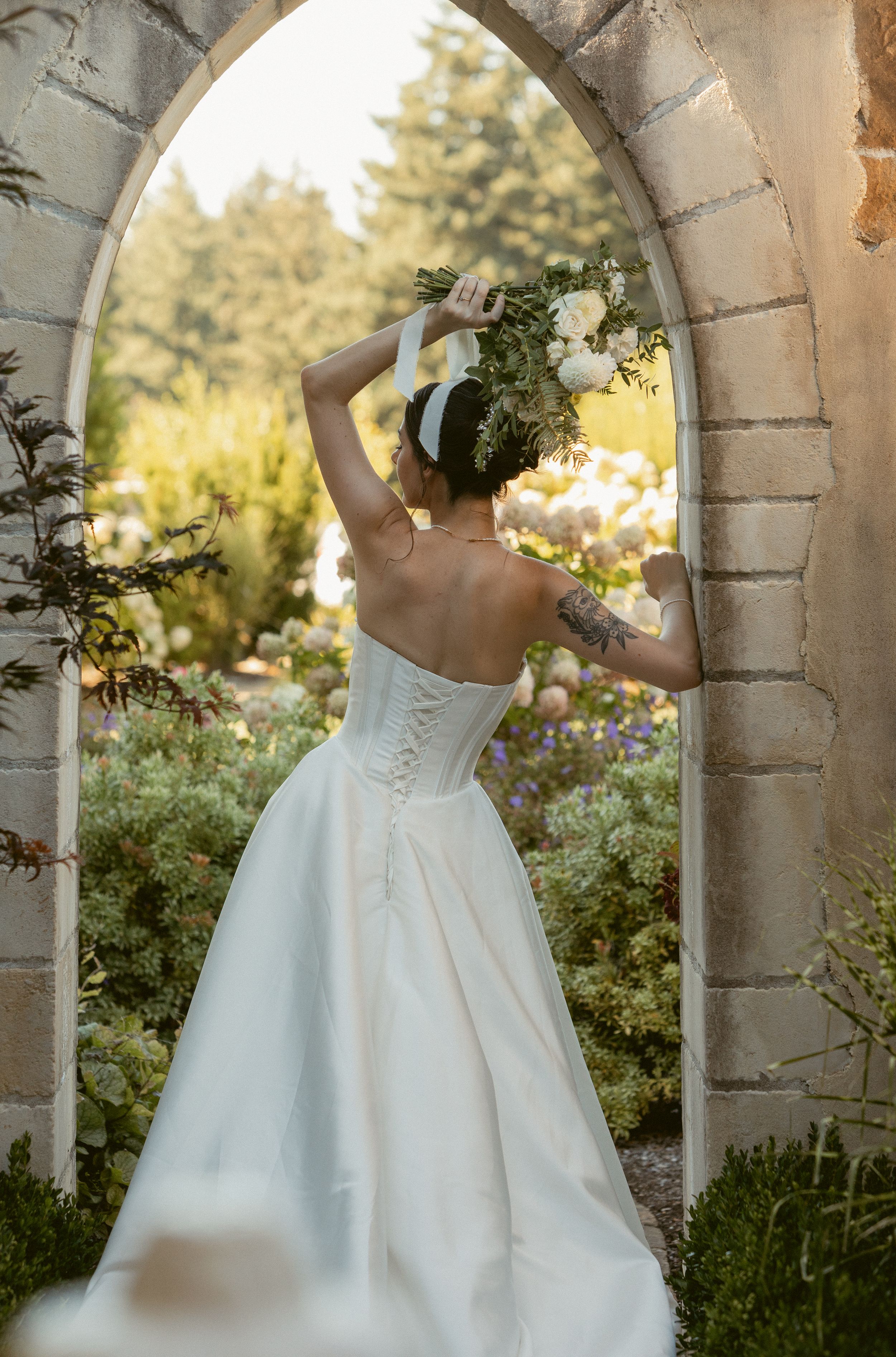 Elegant white wedding dress photographed through stone archway in garden setting at golden hour.