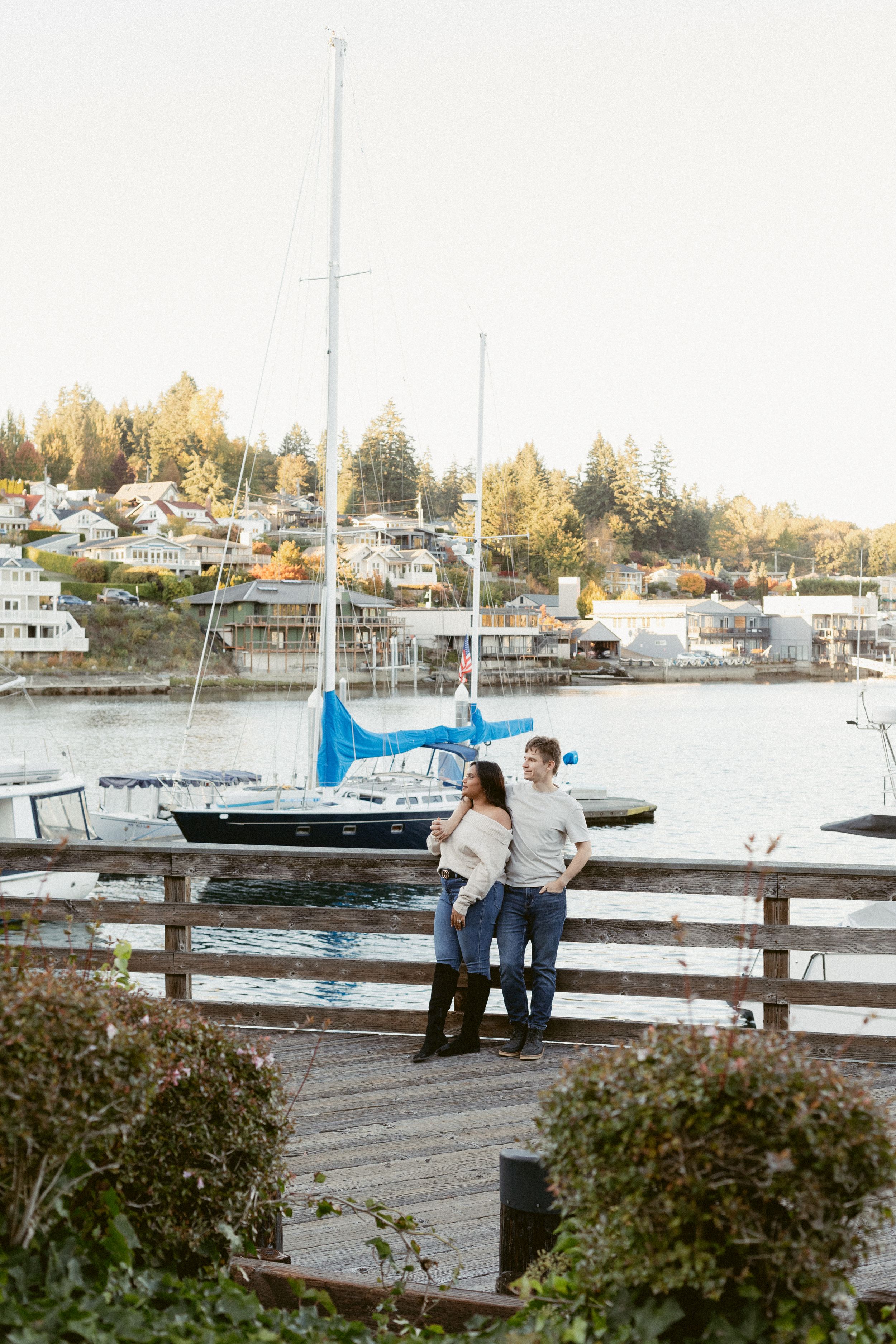 A couple stands together on a wooden dock overlooking a marina with boats and waterfront homes in the background.