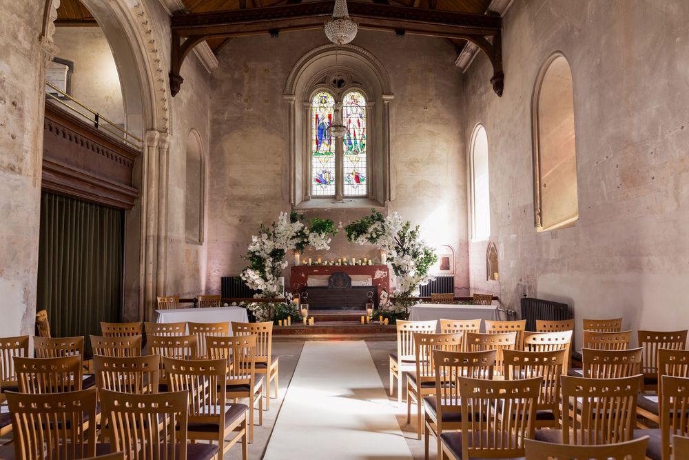 Panoramic view of a historic church interior with wooden chairs arranged in rows and sunlight streaming through stained glass windows.