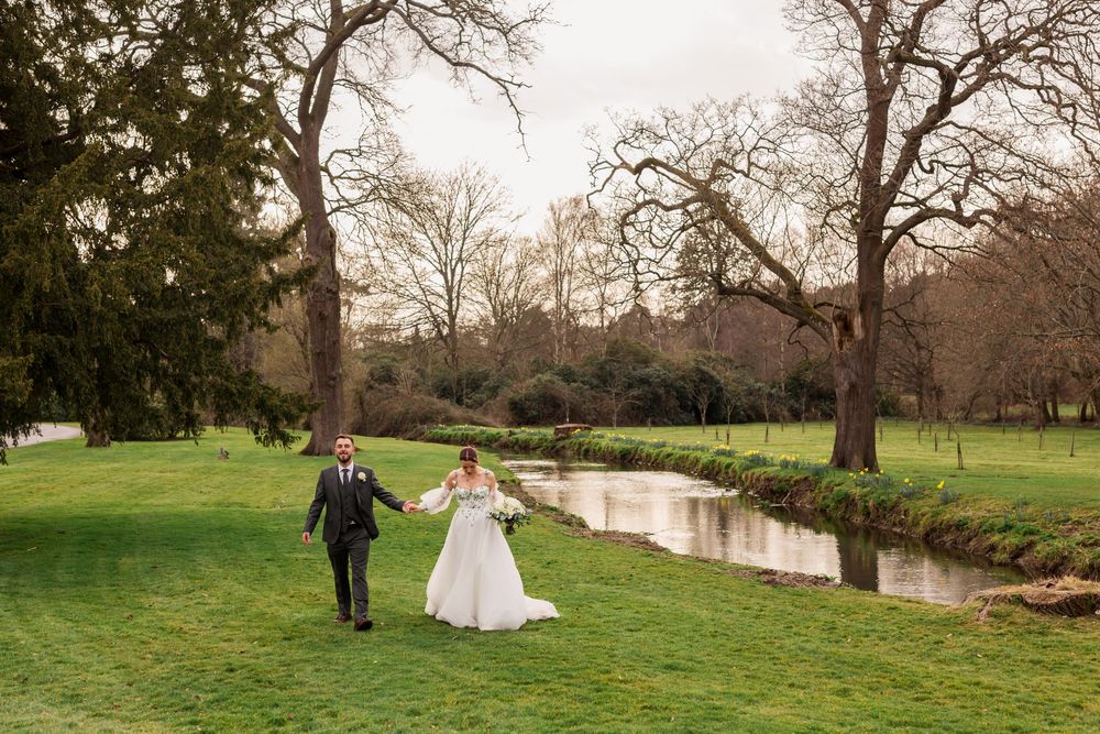 A couple in wedding attire walks together through a scenic park landscape at The Elvetham with tall trees and a winding stream.