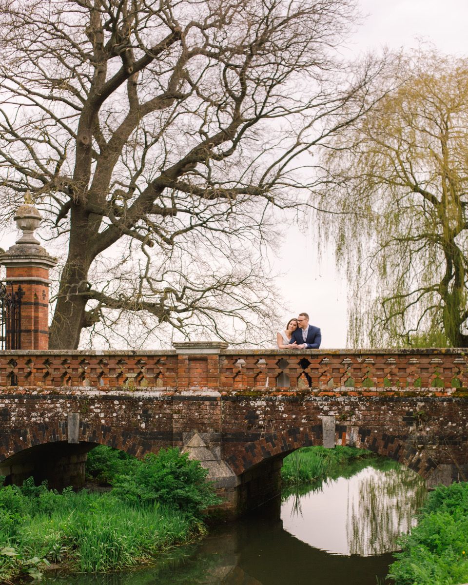 A romantic couple stands on an old brick bridge at The Elvetham overlooking a tranquil stream with bare winter trees in the background.