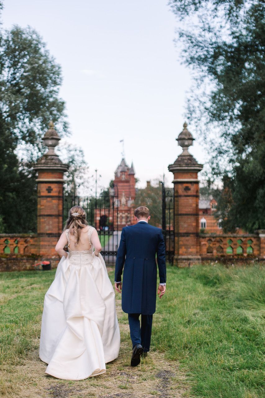 A couple in wedding attire walks toward ornate brick gates and The Elvetham building in the distance.