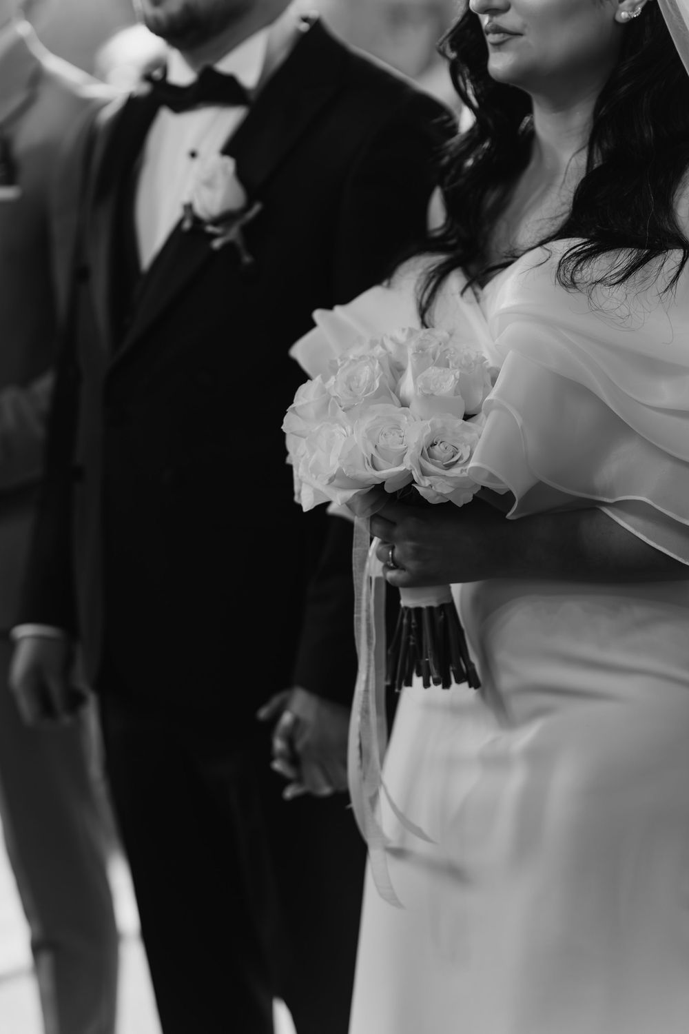 Black and white photo of a wedding couple, focusing on the bride's white rose bouquet and elegant dress details.