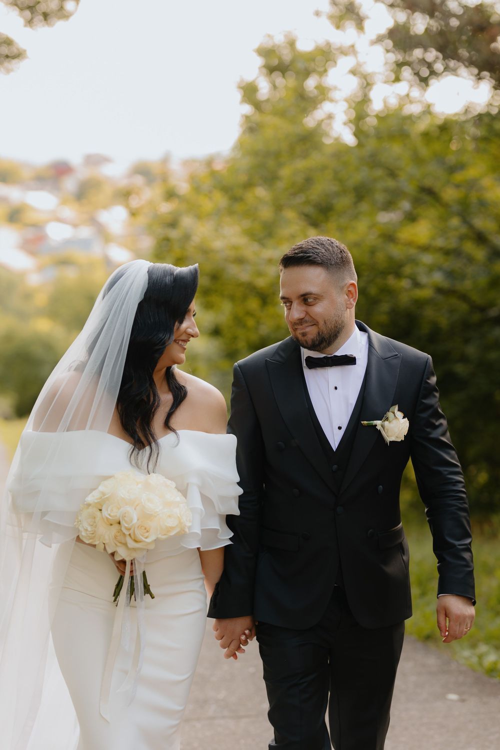 A romantic wedding couple walking together outdoors with the bride holding a cream-colored bouquet.