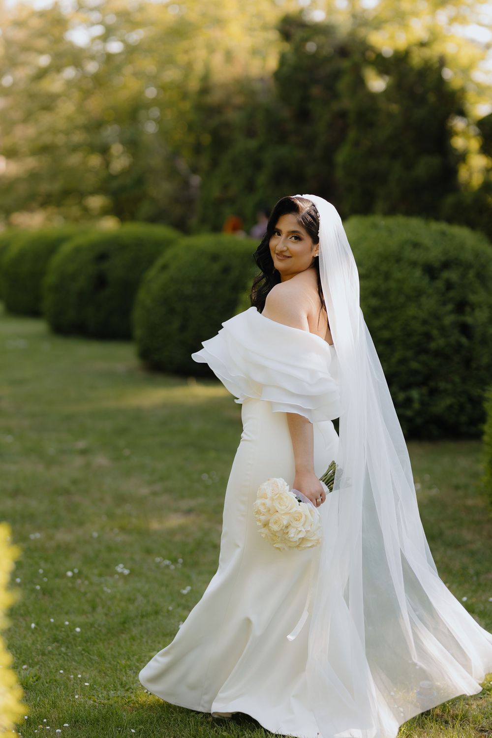 Bride in flowing white wedding dress and veil poses gracefully in a sunlit garden setting.