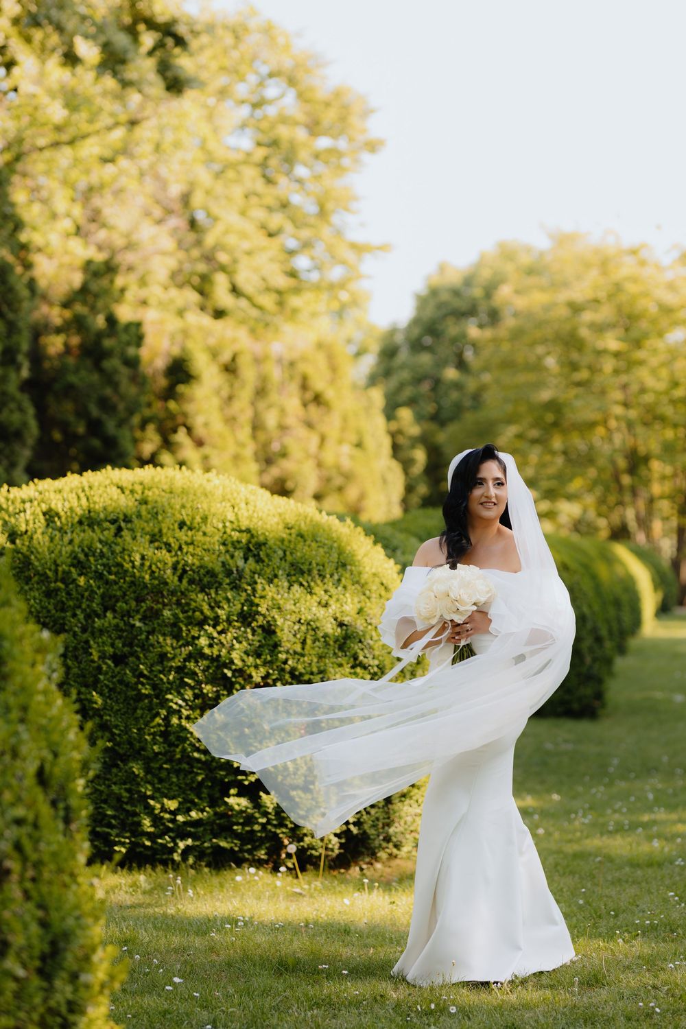 A bride in a flowing white dress twirls in a sunlit garden setting with her veil catching the breeze.