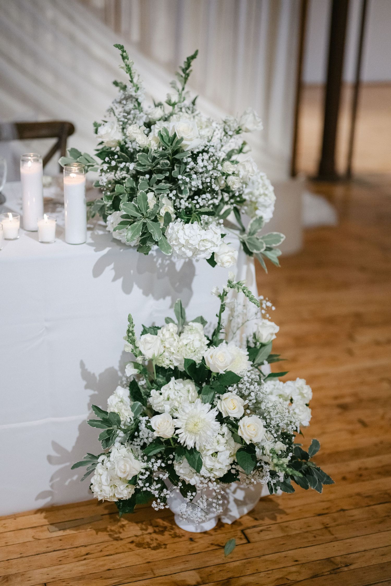 Elegant wedding reception tables with white floral centerpieces, crystal chandeliers, and soft lighting in a banquet hall.