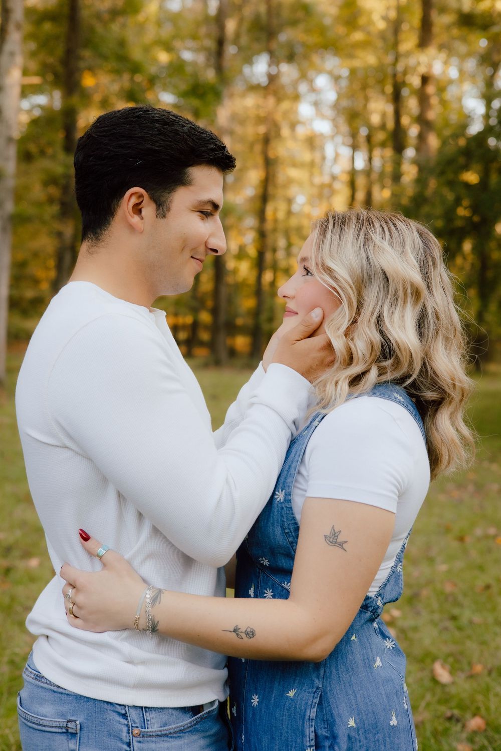 Romantic couple embraces while wearing coordinating white tops and denim in golden fall woods.