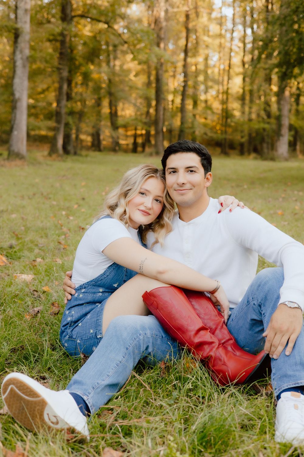 Couple relaxes on grass in casual attire during autumn photoshoot with golden trees in background.
