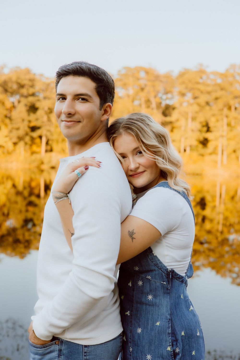 Romantic couple embraces by a serene lake during golden hour with trees in the background.