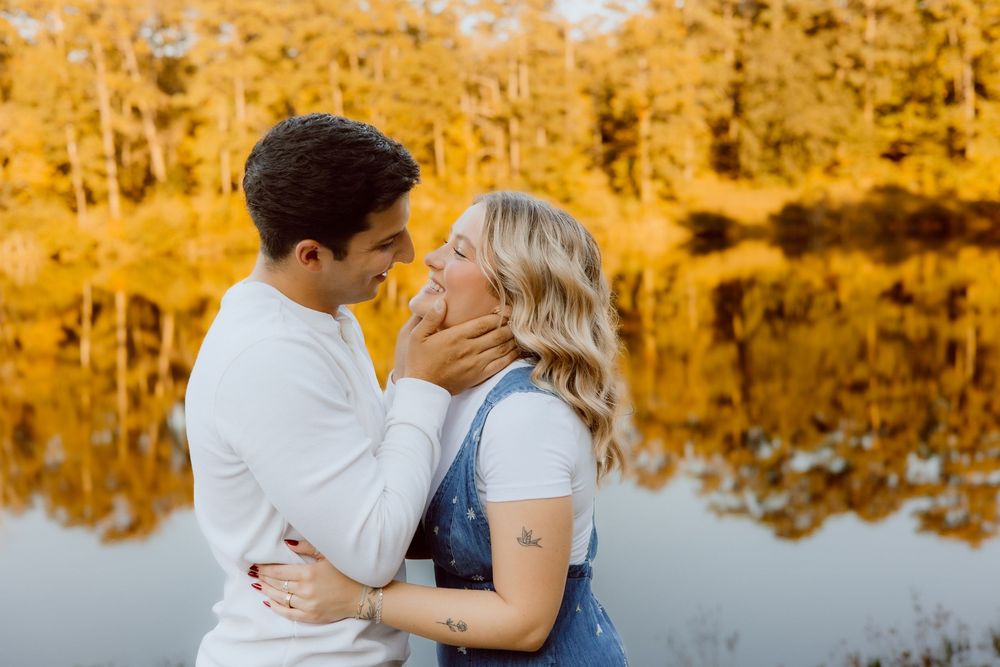 A couple shares a romantic kiss by a lake surrounded by golden autumn trees reflecting in the water.