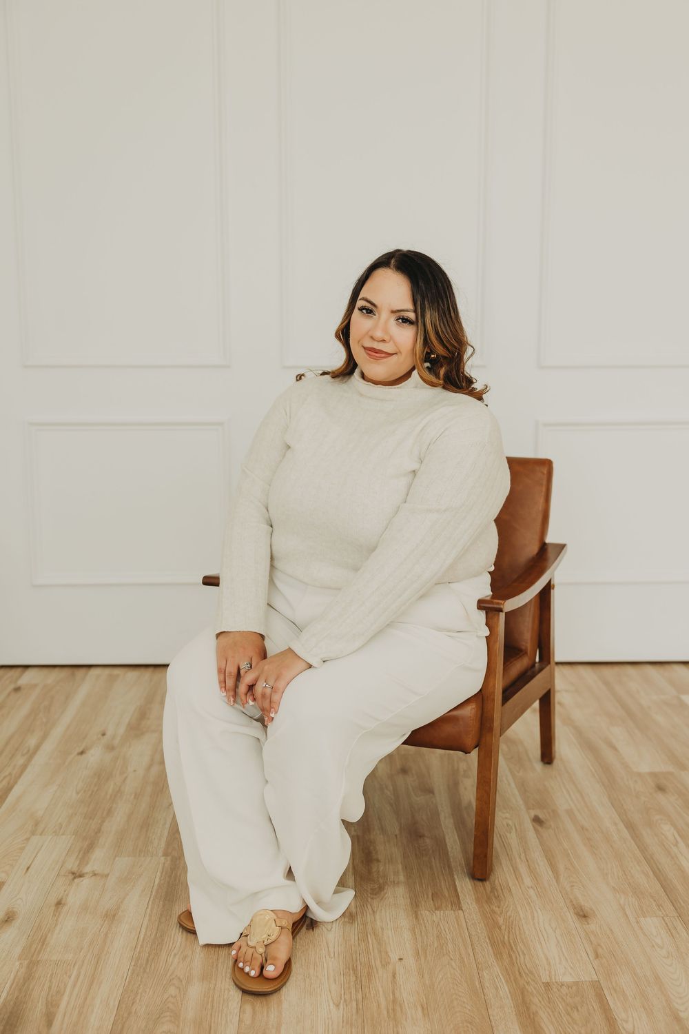 A person wearing a cozy cream-colored sweater and pants lounges in a wooden chair on light hardwood flooring.