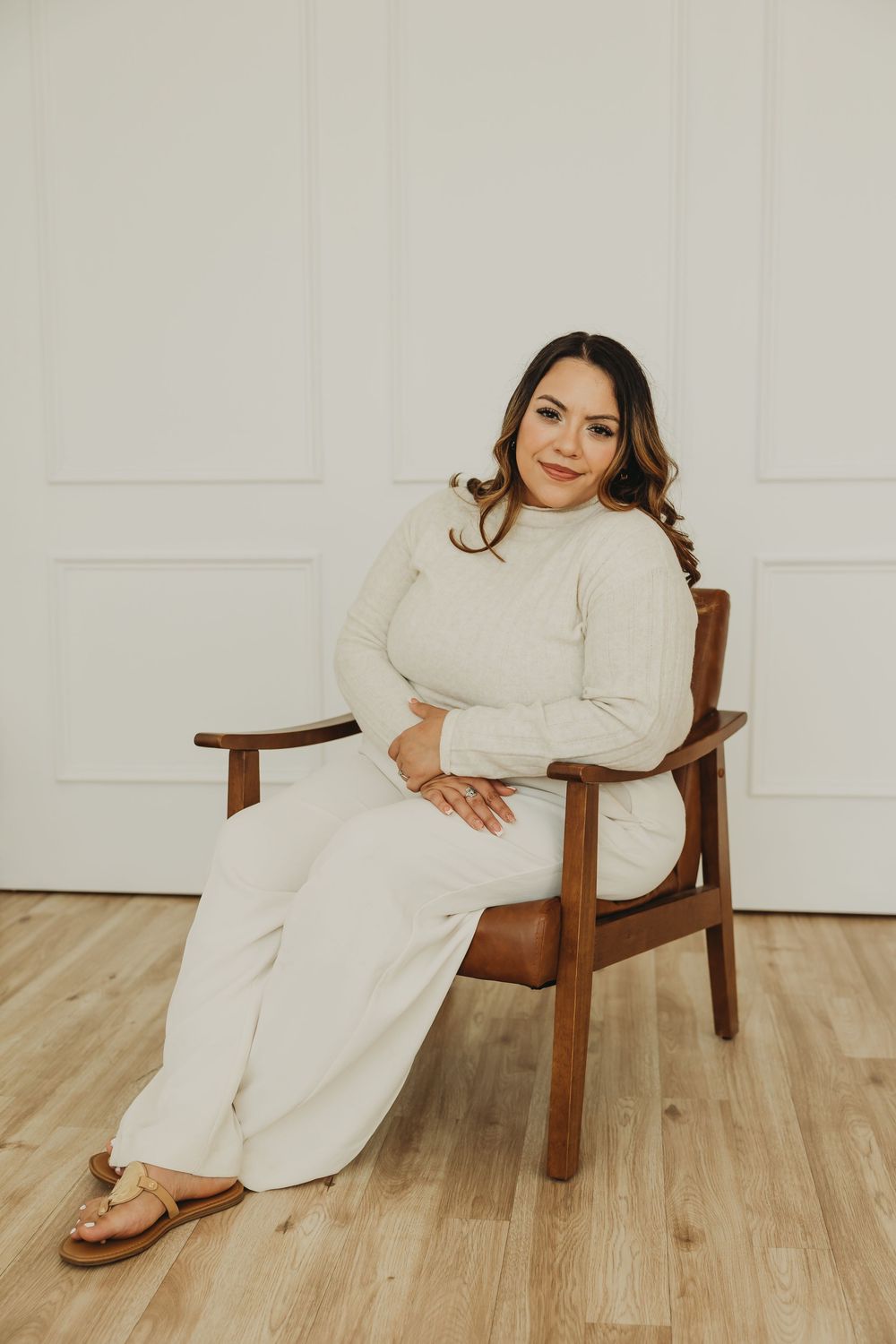 A person in cream-colored loungewear relaxes in a wooden armchair against a white wall.