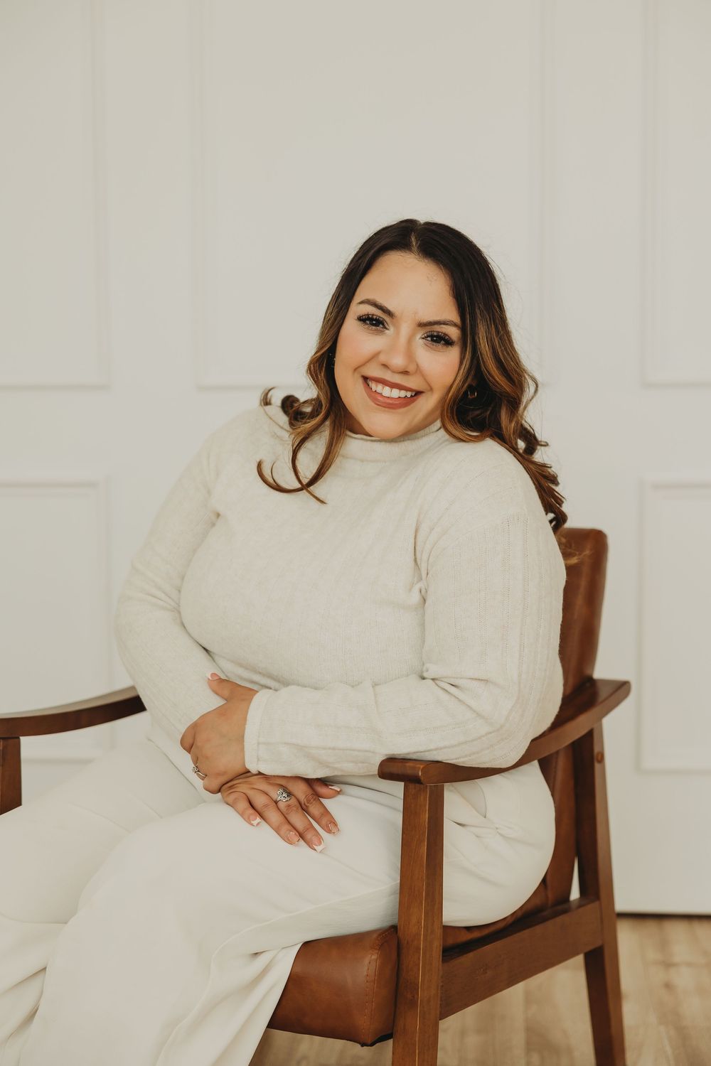 A person in a cozy cream sweater sits comfortably in a wooden armchair against a white wall.