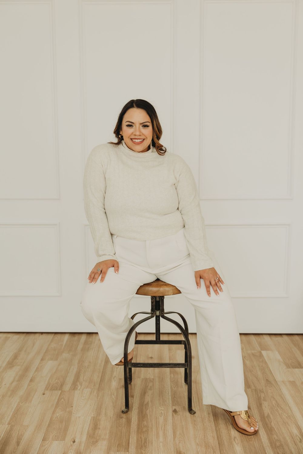 A person wearing a cozy cream turtleneck sweater and matching pants poses on a wooden stool against a white wall.