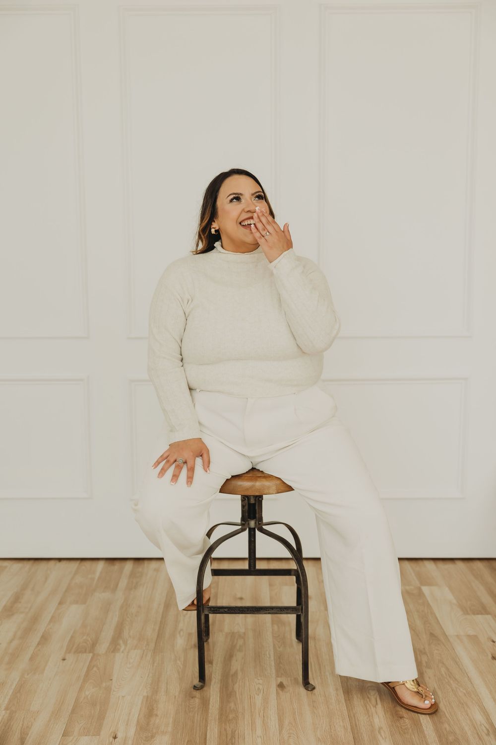 A figure in a white sweater and pants poses expressively on a wooden stool against a light colored wall.