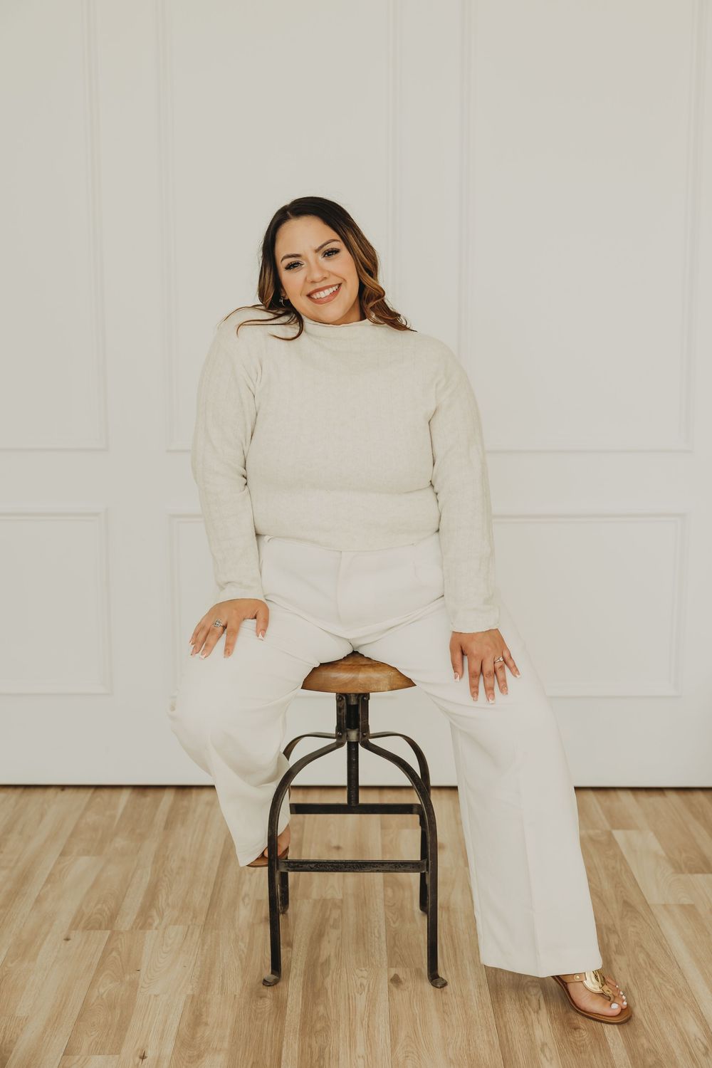 Person in white outfit sits on black metal stool against white wall in minimalist studio setting.