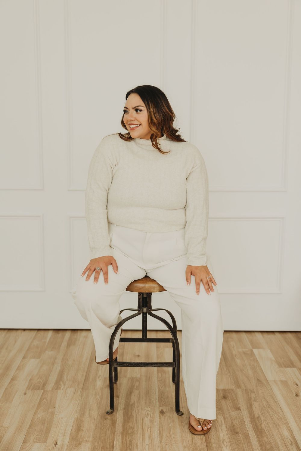 Someone wearing a cozy cream turtleneck sweater and matching pants sits on a wooden stool against a white wall.