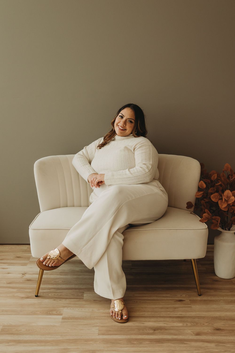 Person in cozy white sweater and pants lounges on vintage cream-colored sofa in minimalist setting with warm lighting.