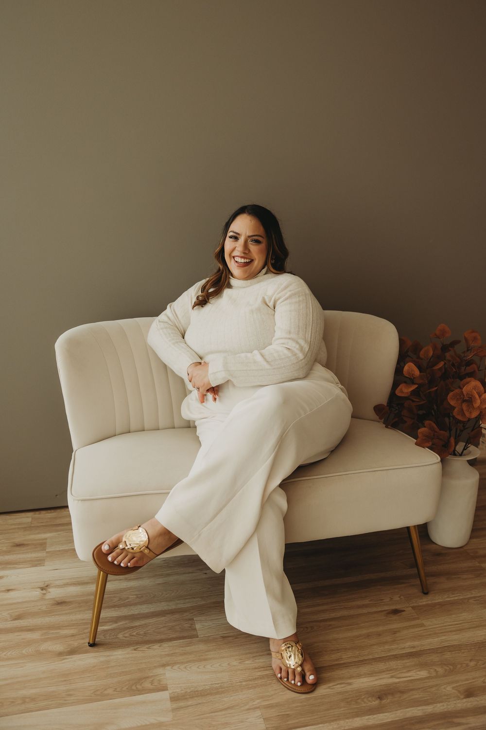 A person in all-white attire relaxes on a light-colored couch with hardwood flooring and autumn decor visible.