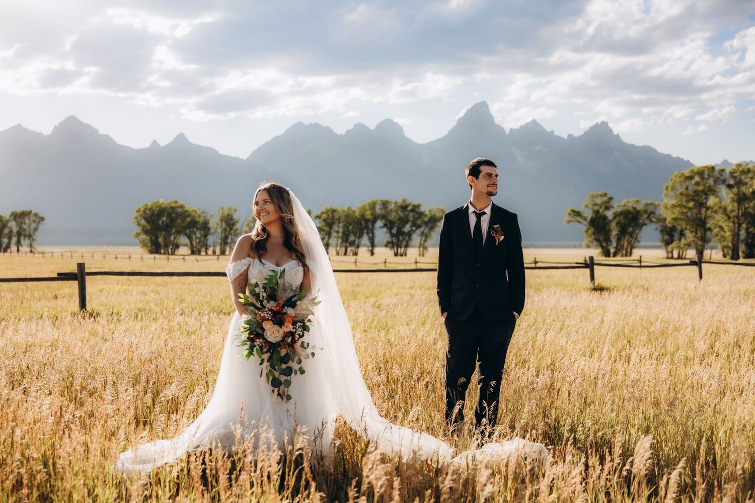 A couple poses in a golden wheat field with majestic mountain peaks rising in the background on their wedding day.