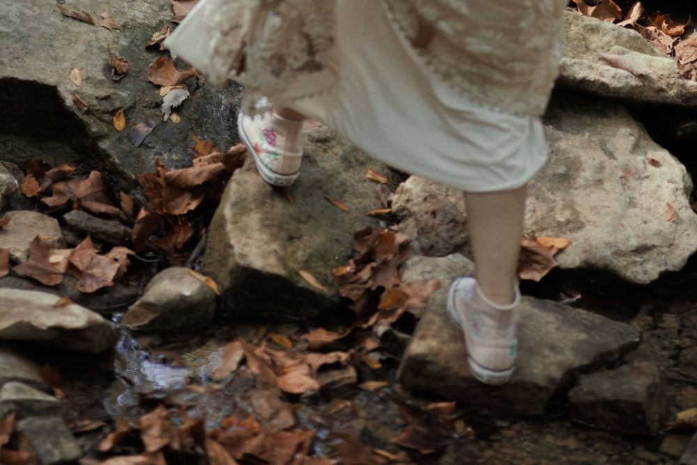 A  bride in a white dress and sneakers steps carefully over mossy rocks and autumn leaves in a forest stream.