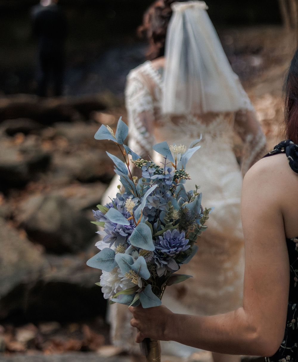 A bridesmaid holds a blue bouquet of succulents and calla lilies with a veiled bride in a lace gown in the background.