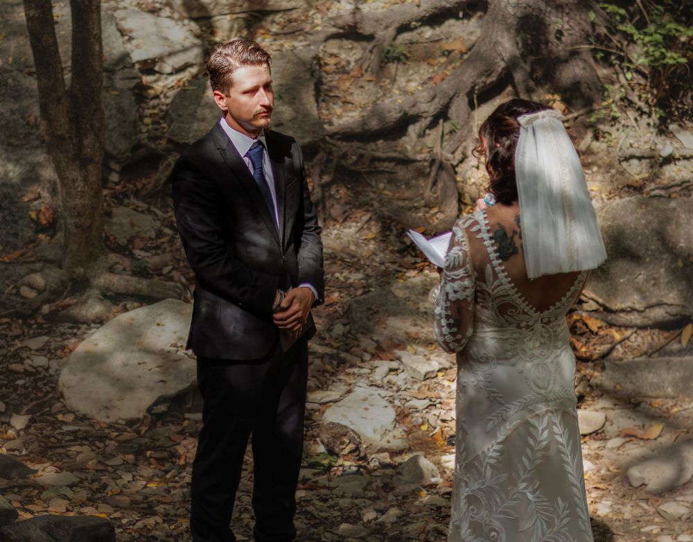 Bride in lace gown and veil reads vows to groom in black suit during intimate outdoor woodland wedding ceremony.