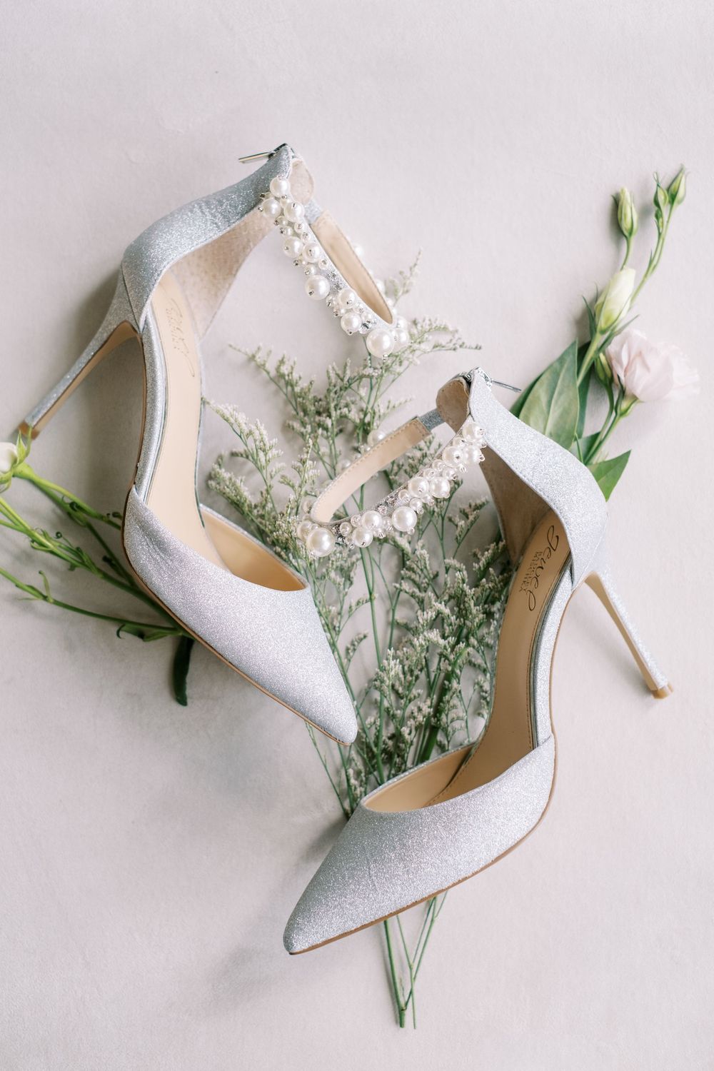 Elegant white glitter bridal heels arranged with delicate white flowers and greenery on light gray background.