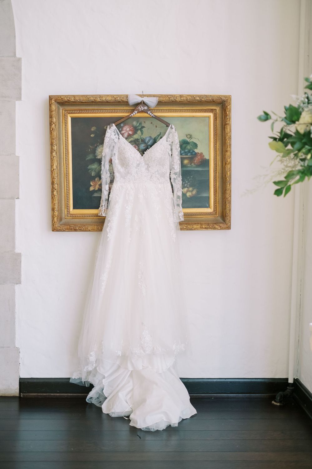 White lace wedding dress hanging on display in front of elegant gold-framed artwork on a white wall.