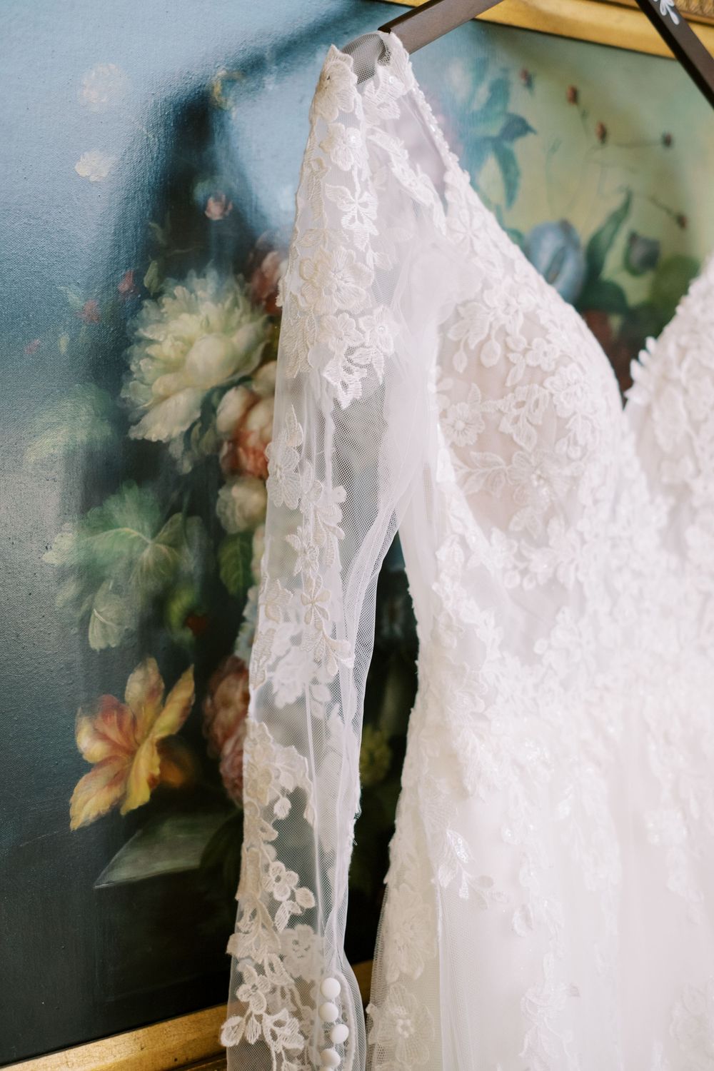 Close-up detail of a delicate white lace wedding dress sleeve hanging against a blue floral wallpaper background.