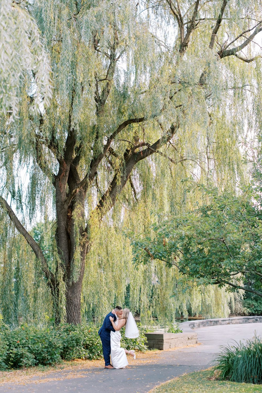 A romantic moment under weeping willow trees beside a tranquil pond in a serene garden setting.