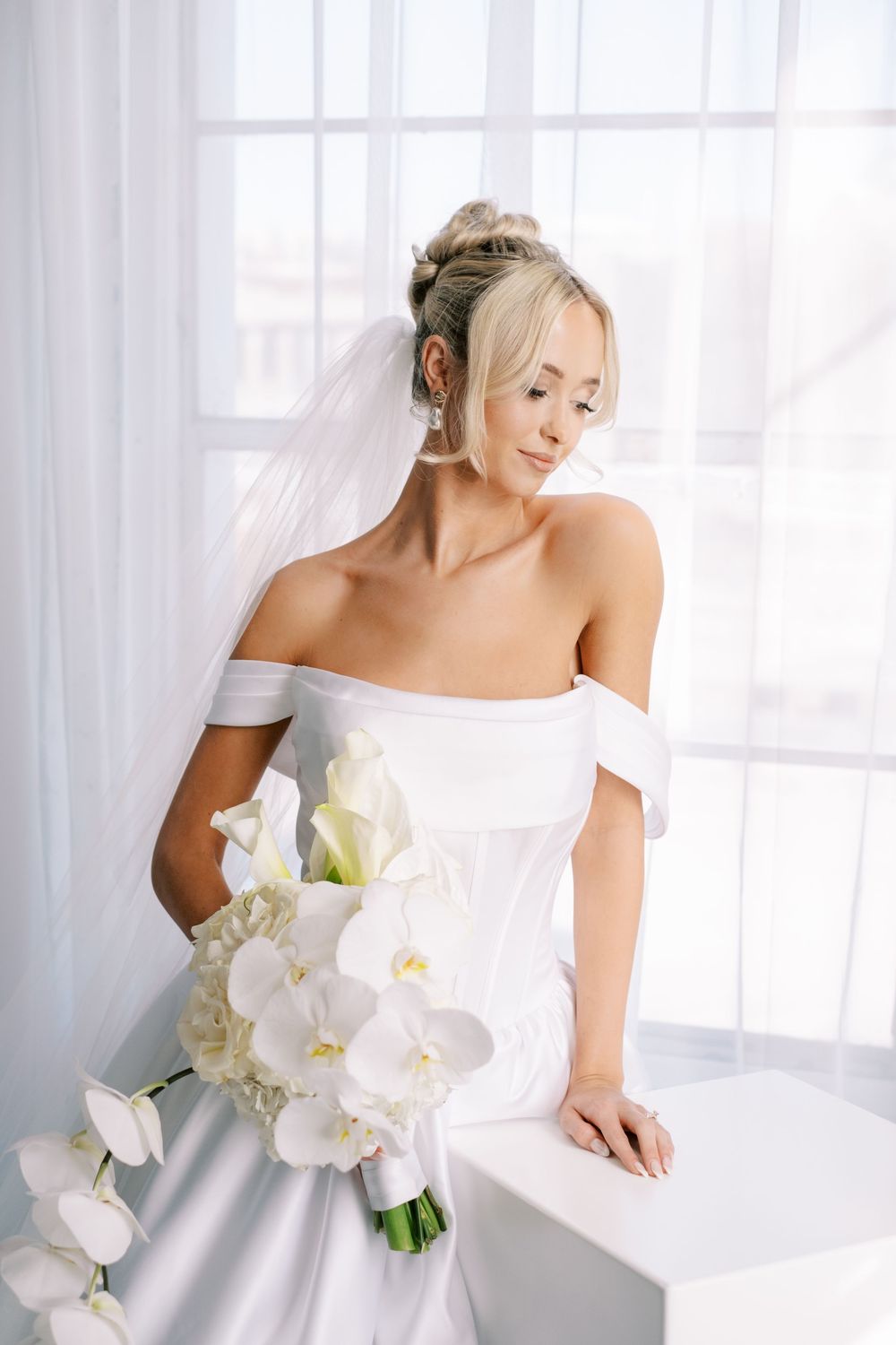 Elegant bridal portraits in white with delicate orchid bouquet against soft window light and sheer curtains.