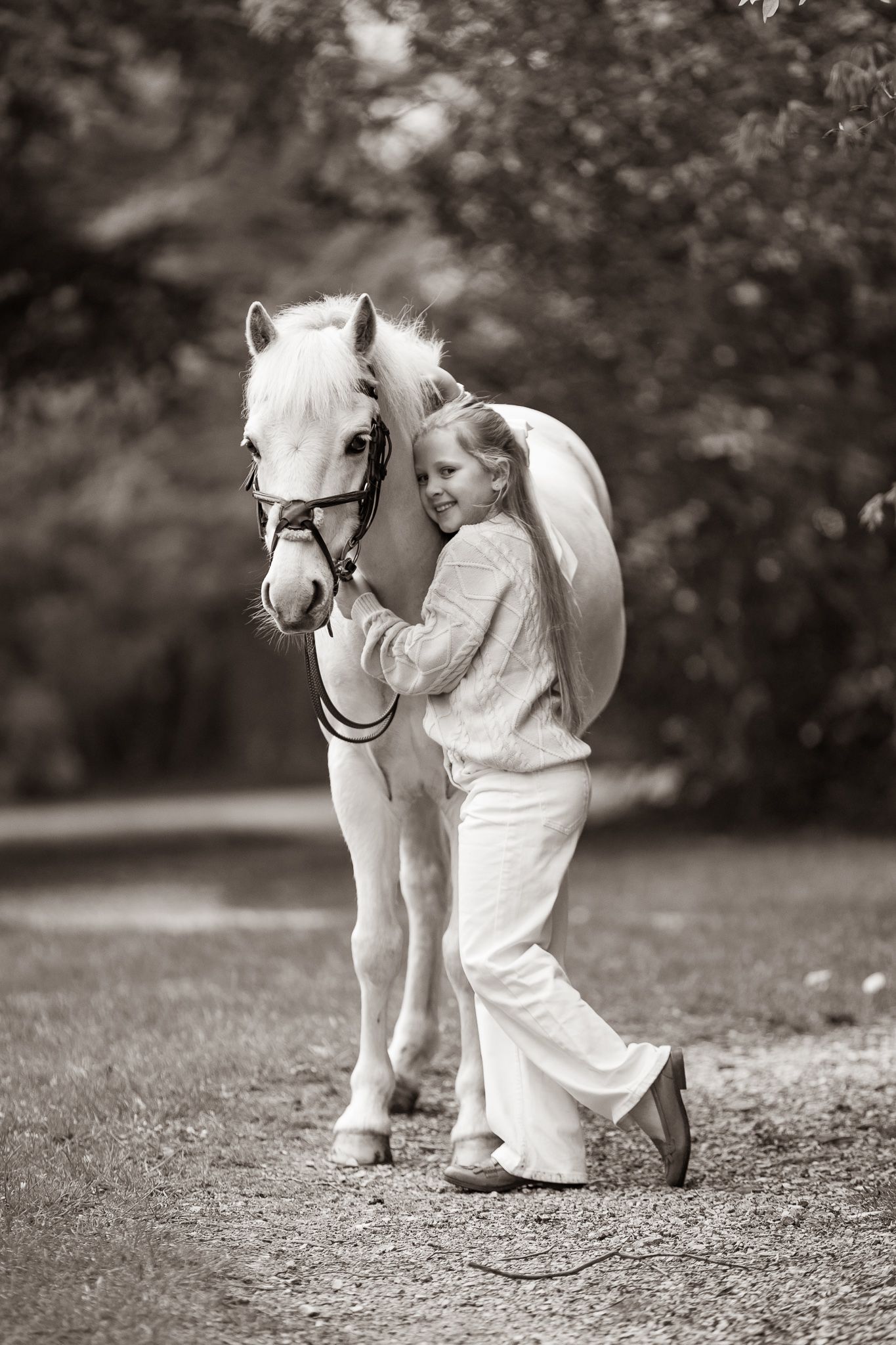 Black and white photo of a smiling girl in white outfit walking with a white pony on a tree-lined path.