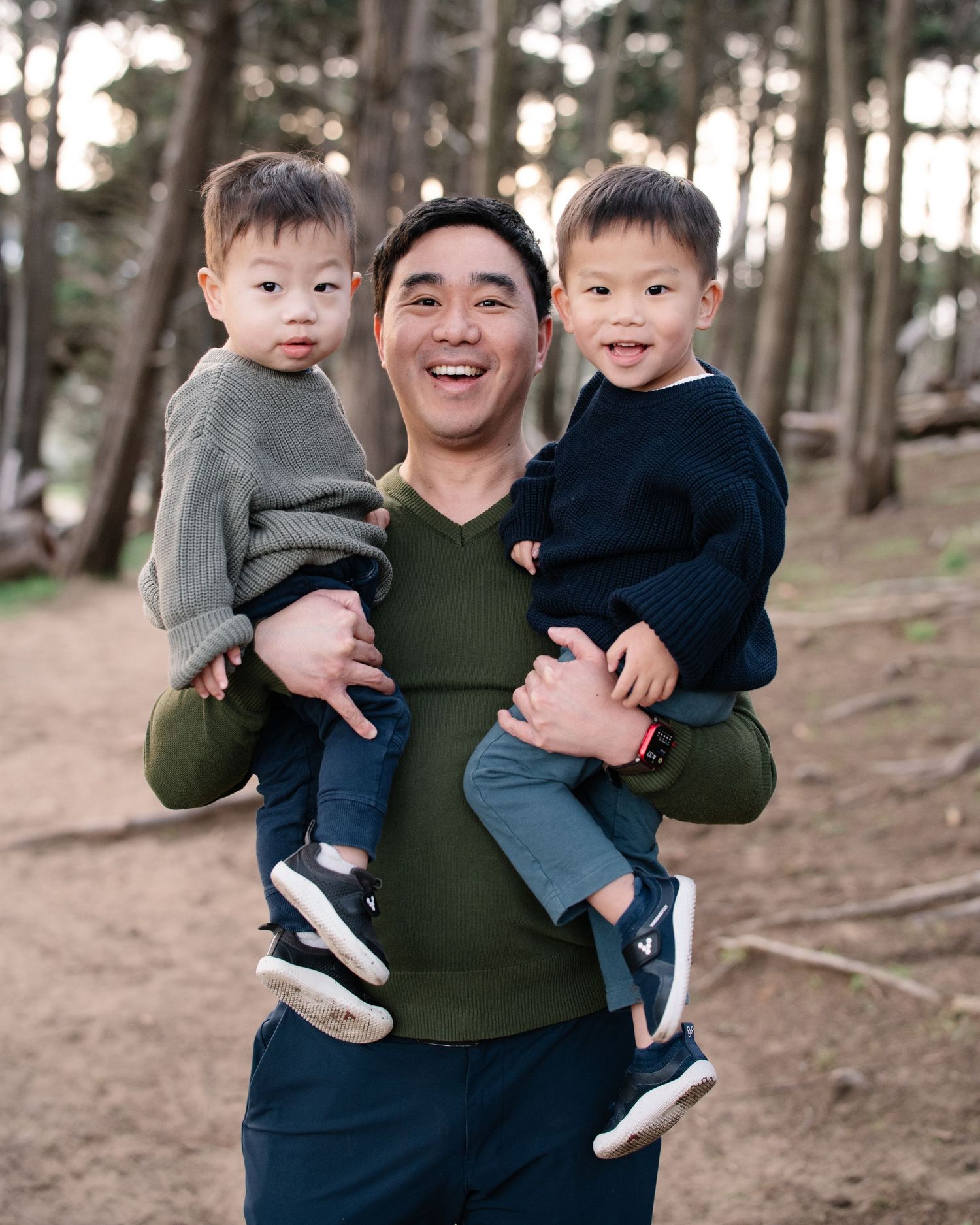 A smiling man holds two young boys in his arms while standing on a wooded trail outdoors.
