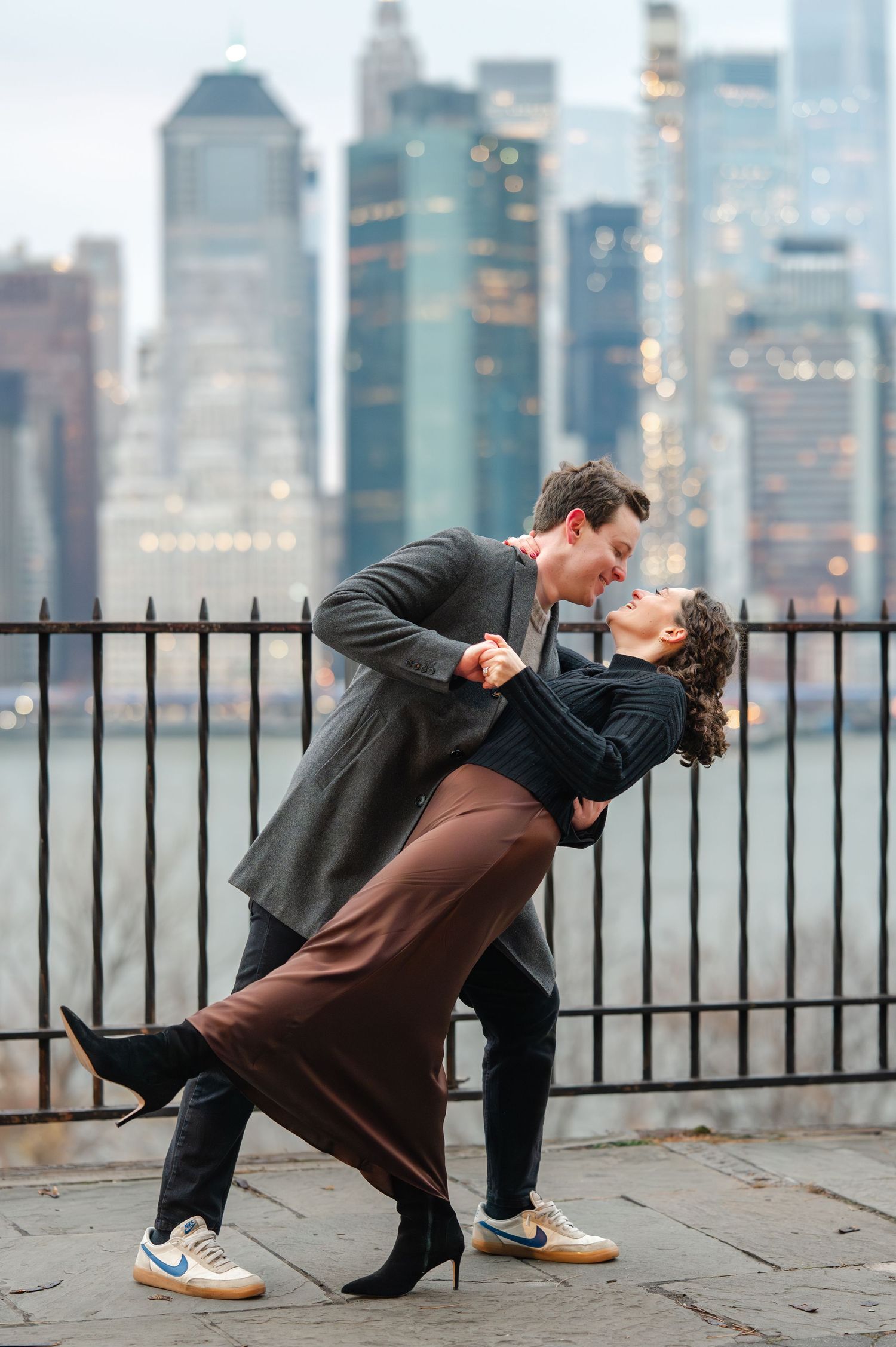 Couple dancing a romantic dip in front of NYC skyline, woman in brown skirt and black heels, man in gray coat.