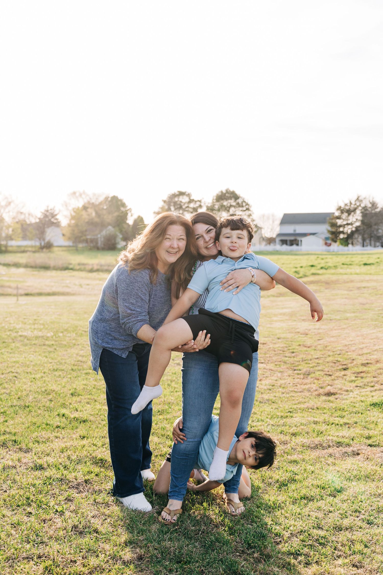 Two women and two children laugh and play together in a sunny green field during a warm family photo session outdoors.