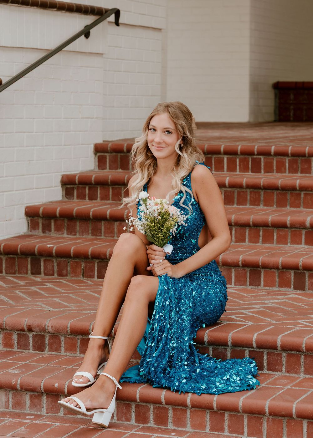 Young woman in teal sequin gown holding white flowers, seated on brick steps for a formal occasion.