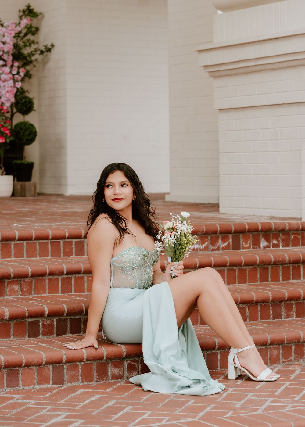 Young woman in mint green gown holding wildflowers, seated on brick steps outdoors.