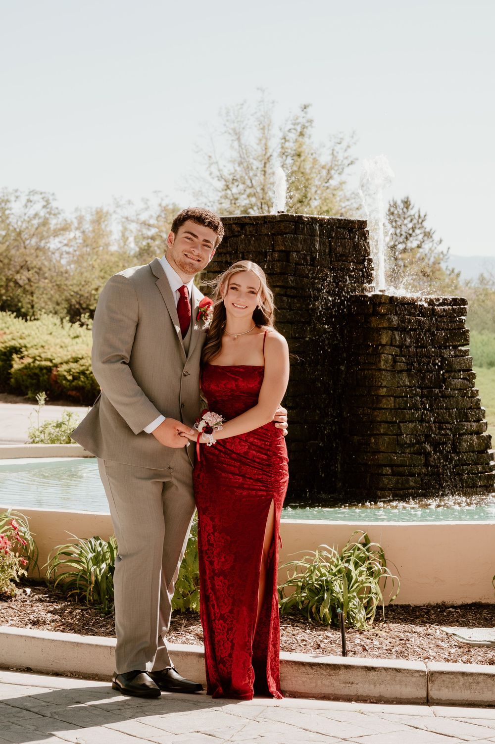 Couple posing in formal wear by fountain, man in tan suit with red tie, woman in red sequin gown.